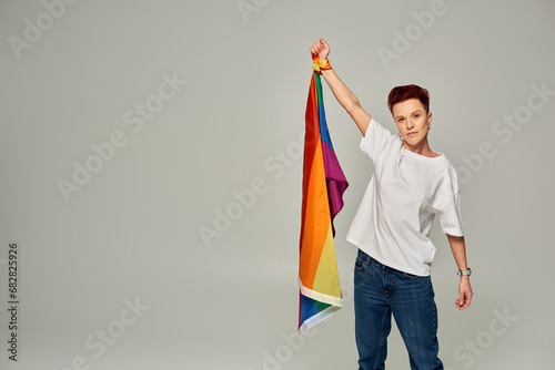 serious redhead bigender person in white t-shirt and jeans standing with LGBT flag on grey backdrop