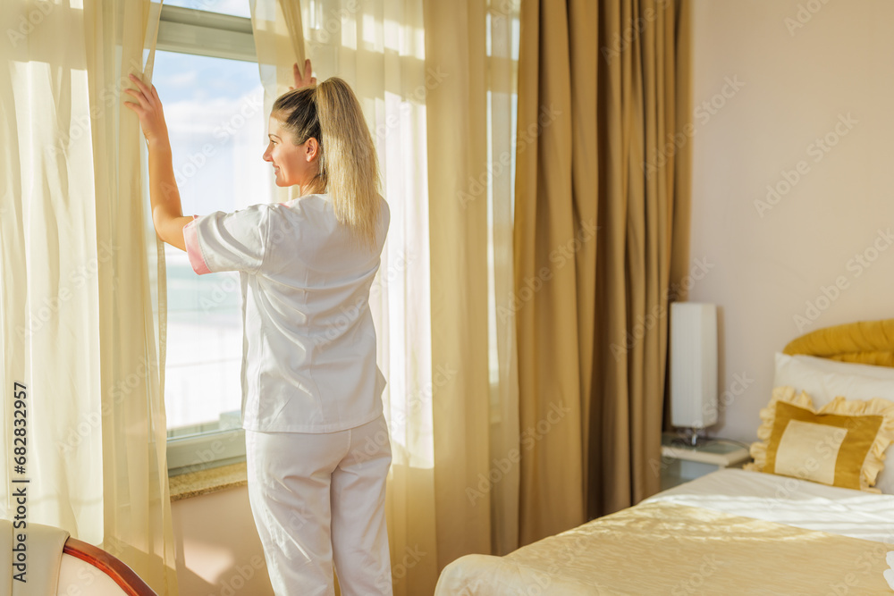 Image of beautiful  hotel maid opening window curtains  in a room.