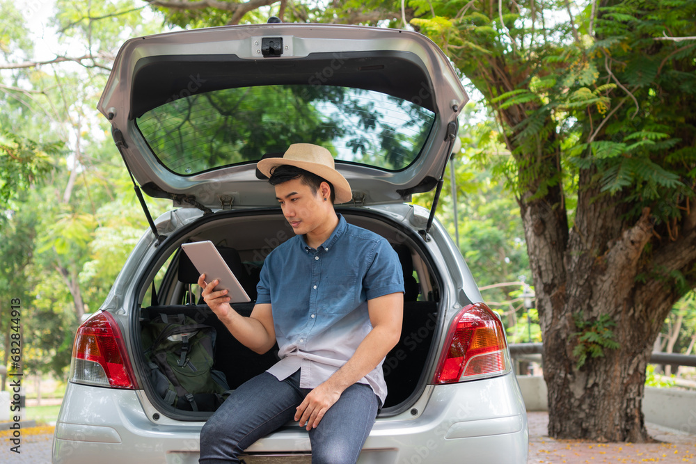 Asian man sitting back of car and holding tables check location and find place to travel. Happy road trip and vacation in summer in the countryside. Concept of journey on leisure, road trip vacation Asian man sitting back of car and holding tables check location and find place to travel. Happy road trip and vacation in summer in the countryside. Concept of journey on leisure, road trip vacation