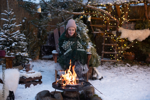 Young attractive woman in winter clothes sitting in a armchair, warming herself by the fire and relaxing outside in winter
