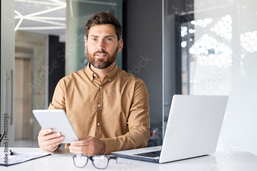 Young and confident male businessman working in the office, sitting at a table with a laptop, using a tablet and looking seriously at the camera