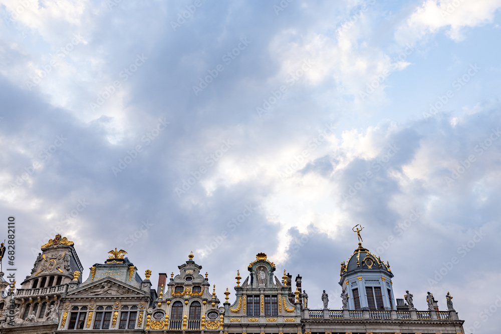 The image captures the grandeur of historic architecture in Brussels against a dynamic sky. The intricate facades and golden statues atop the buildings exude opulence and the artistry of classical