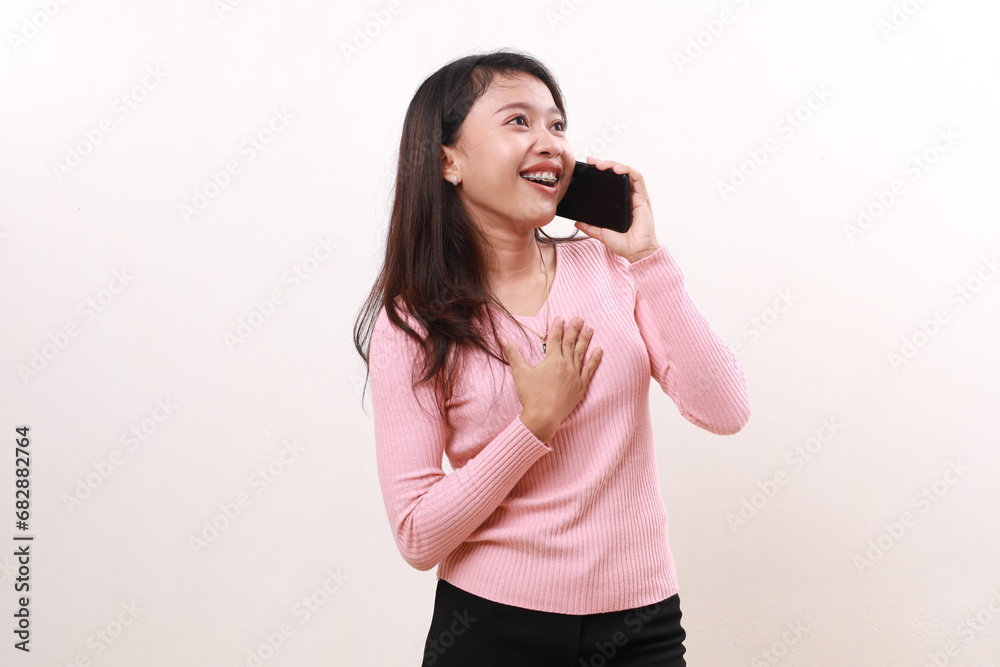 Happy relieved young asian girl standing holding her chest while talking on the phone. Isolated on white background Happy relieved young asian girl standing holding her chest while talking on the phone. Isolated on white background