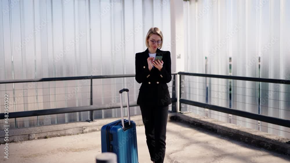 Young businesswoman in a black coat uses mobile phone, standing by her blue suitcase at an airport. Female traveler holding smartphone device enjoying modern technology. Checking online banking app Young businesswoman in a black coat uses mobile phone, standing by her blue suitcase at an airport. Female traveler holding smartphone device enjoying modern technology. Checking online banking app