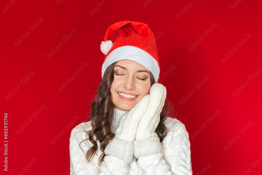 a young pretty woman in a Santa Claus hat and a knitted white sweater holds her hands in white knitted mittens under her cheek, closing her eyes and smiling. isolated on a red background a young pretty woman in a Santa Claus hat and a knitted white sweater holds her hands in white knitted mittens under her cheek, closing her eyes and smiling. isolated on a red background