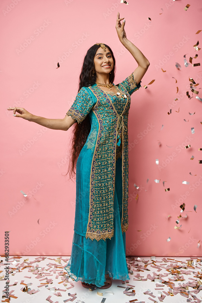 vertical shot of young indian woman smiling at camera gesturing while dancing under confetti rain