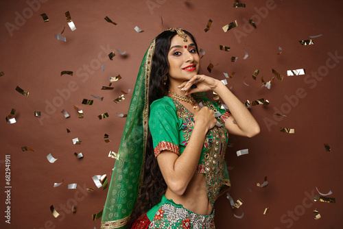 pretty indian woman with bindi dot and green veil posing under confetti rain with hand under chin photo