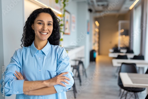 Portrait of young joyful successful Latin American small business owner, slim boss smiling and looking at camera with crossed arms, businesswoman at entrance to cafe restaurant, inviting visitors.