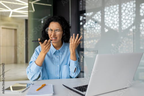 Annoyed angry businesswoman at workplace inside office, female worker shouting recording audio message using app on phone, woman with curly hair.