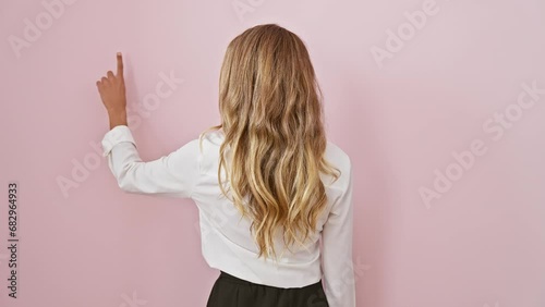 Striking young blonde woman posed for a captivating back view shot on a pink isolated background, wearing a shirt as she stands, pointing forwards with efficiency and strong air of leadership. photo