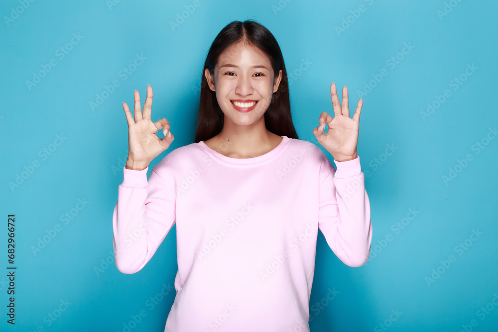 Beautiful young woman posing with OK hand gesture, Portrait of a beautiful young woman in a light blue background, happy and smile, posting in stand position. Beautiful young woman posing with OK hand gesture, Portrait of a beautiful young woman in a light blue background, happy and smile, posting in stand position.