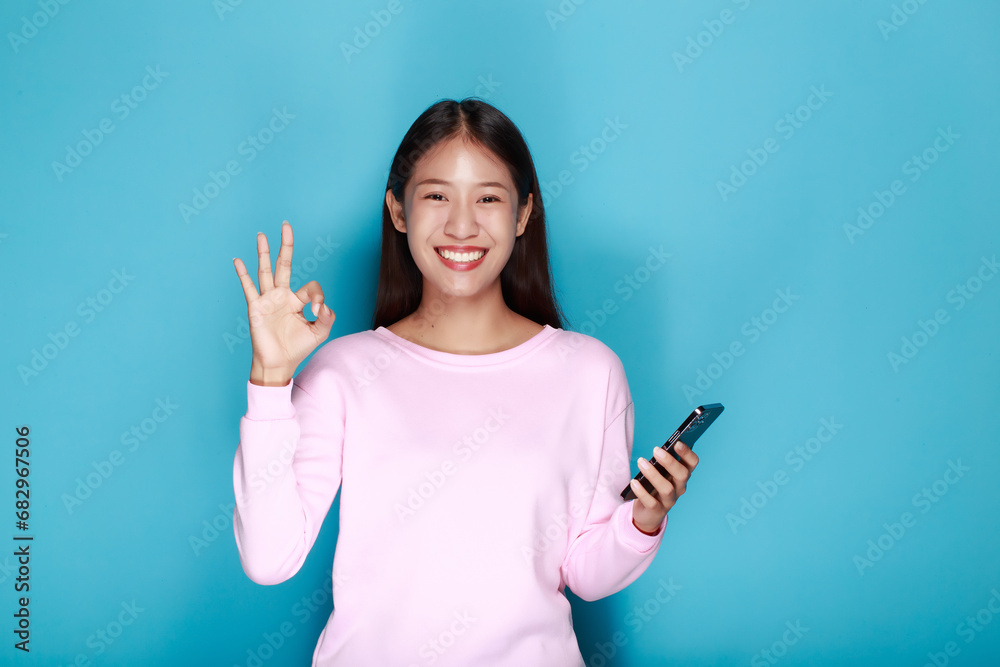 Young woman poses with OK hand while holding a cell phone in the other hand, Portrait of a beautiful young woman in a light blue background, happy and smile, posting in stand position. Young woman poses with OK hand while holding a cell phone in the other hand, Portrait of a beautiful young woman in a light blue background, happy and smile, posting in stand position.