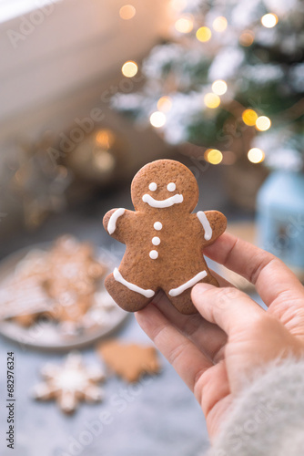 A baked of cut out Christmas sugar biscuit in a gingerbread man shape while hand holding it. With various shapes of cooked cookies and cozy festive ambiance lighting bokeh in background