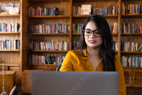 A beautiful young girl student wearing glasses or spectacles and studying in school or collage library or working in corporate office using laptop searching for information on internet or books