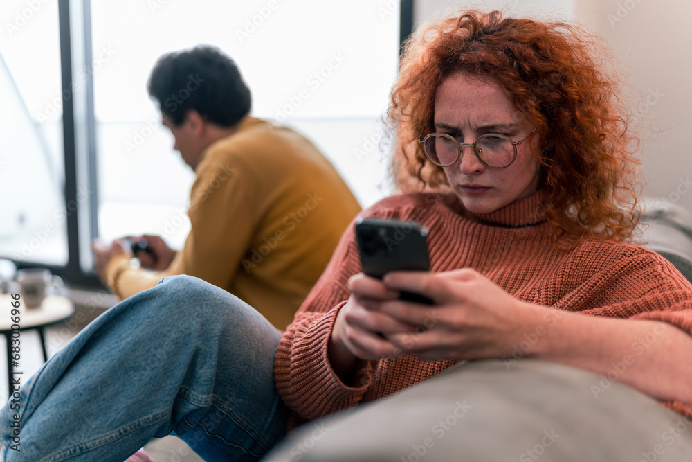 An interracial couple sitting on the couch, she is looking at her phone and is very serious and mad, he is playing a game and is concentrating in the background An interracial couple sitting on the couch, she is looking at her phone and is very serious and mad, he is playing a game and is concentrating in the background