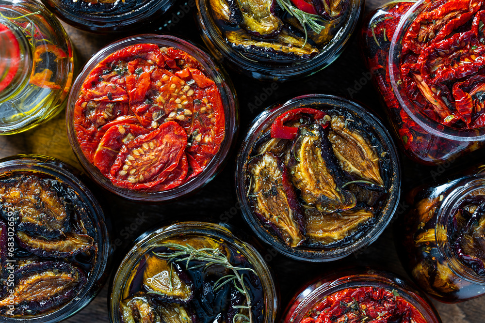 Sun-dried red tomatoes and plums with garlic, green rosemary, red chili pepper, olive oil and spices in a glass jars on a wooden table. Rustic style, top view, closeup - obrazy, fototapety, plakaty 