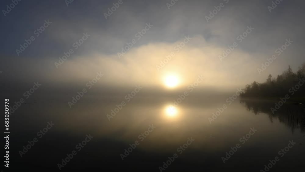 Morning fog drifts across a calm lake with the rising sun creating a bright circle near the center of the clip. A point of land juts into the lake on the right side. The calm water reflects the treed Morning fog drifts across a calm lake with the rising sun creating a bright circle near the center of the clip. A point of land juts into the lake on the right side. The calm water reflects the treed