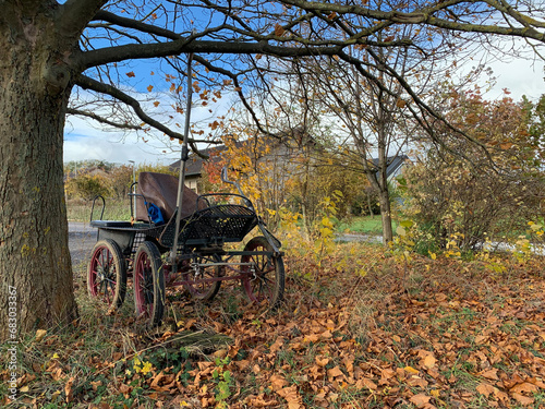 An old open single-seater four-wheeled horse-drawn carriage without a horse. A tarantass or carriage stands under a tree in autumn among fallen leaves in sunny weather in a German village in Germany.