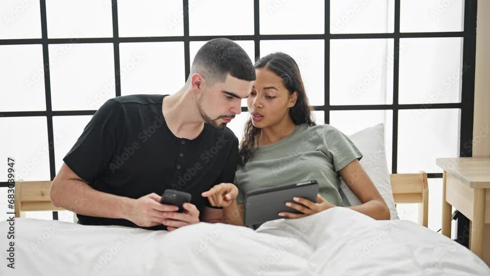 Beautiful couple sitting together on a cozy bed in the bedroom, immersed in their smartphone and touchpad, basking in the relaxed morning at home Beautiful couple sitting together on a cozy bed in the bedroom, immersed in their smartphone and touchpad, basking in the relaxed morning at home