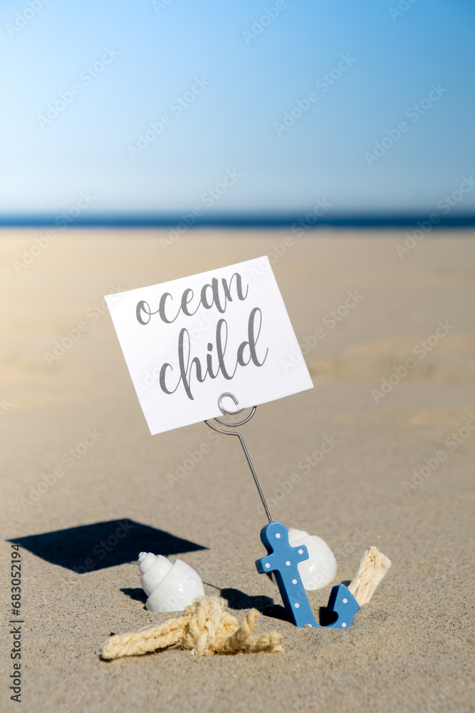 Obraz premium OCEAN CHILD text on paper greeting card in anchor paper holder and starfish seashell summer vacation decor. Sandy beach sun coast. Holiday concept postcard. Getting away Travel