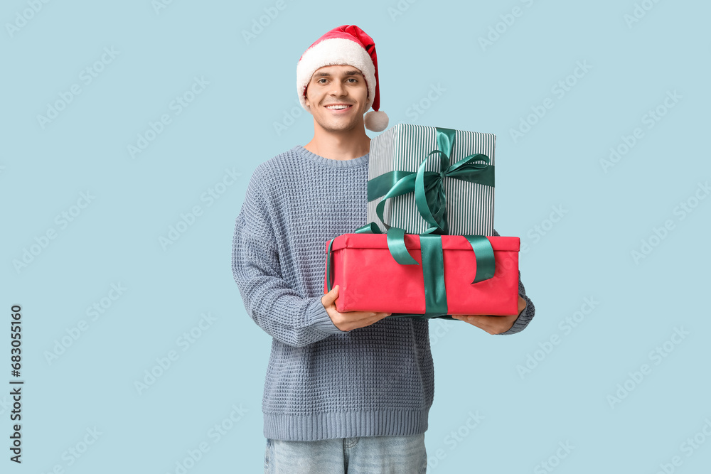 Handsome young man in Santa hat holding Christmas gifts on blue background Handsome young man in Santa hat holding Christmas gifts on blue background