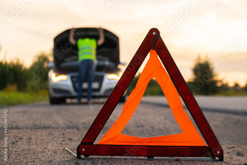 Unrecognizable sad driver in reflective vest. Male driver standing near broken car with open up hood. Red triangle to warn other road users of car breakdown or engine failure stop at countryside 