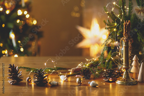 Stylish candle, golden lights, pine cones and ornaments on wooden table against stylish decorated christmas tree with festive illumination. Atmospheric winter holidays at festive rustic home