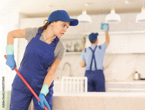 Positive girl in blue overall - employee of cleaning company can use mop to floor kitchen and together with guy partner performs order photo