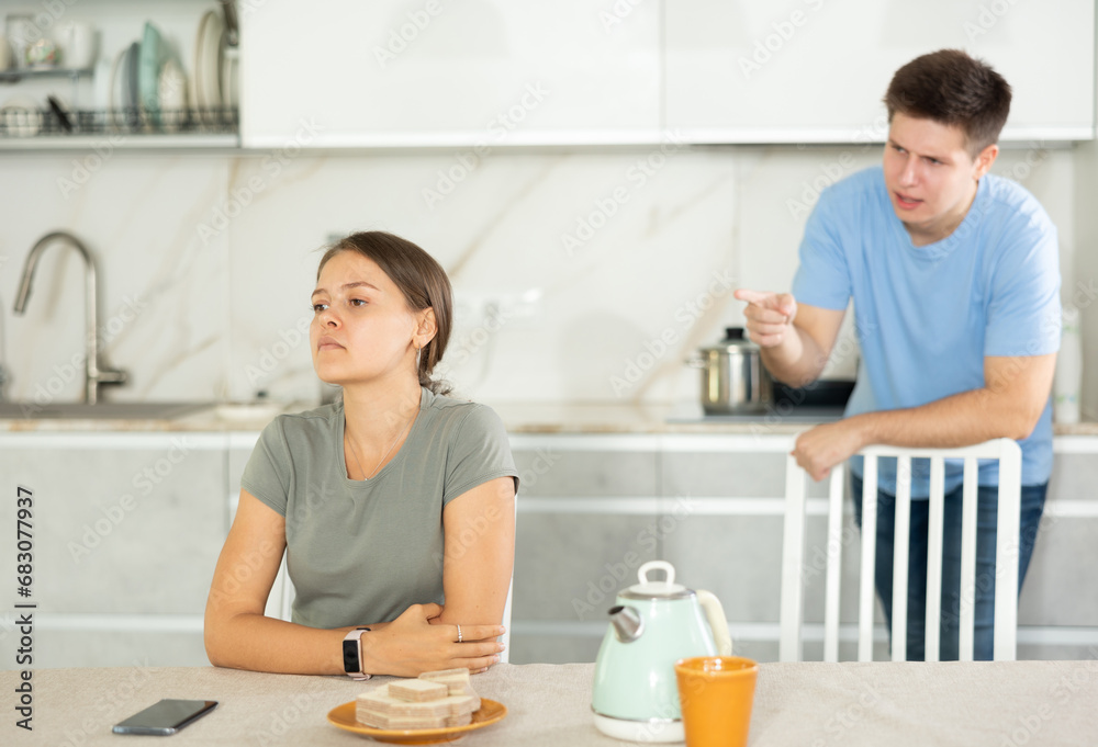 Sad young girl sitting at the table while her boyfriend is quarreling with her standing behind in the kitchen