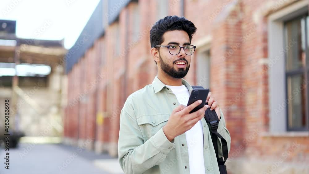 A handsome student with a backpack uses a smartphone while walking around the campus space near the university building. Happy smiling male chatting online, browsing social media or reading messages