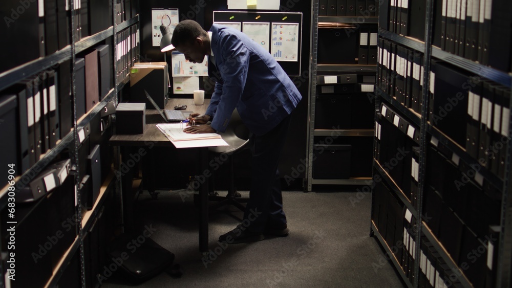 African American policeman carefully analyzes material, does records searches, and collects data from shelves and laptop. Male police officer standing, looking through case files on desk in office. African American policeman carefully analyzes material, does records searches, and collects data from shelves and laptop. Male police officer standing, looking through case files on desk in office.