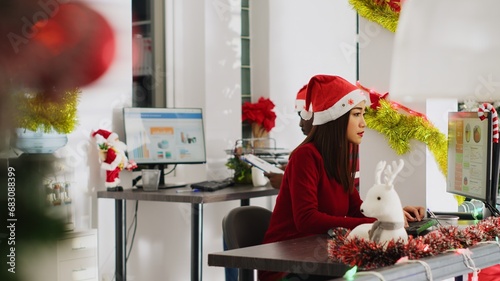 Revealing shot of hardworking asian employee working in Christmas ornate office. Diligent worker looking over financial figures at computer desk in diverse xmas ornate workplace