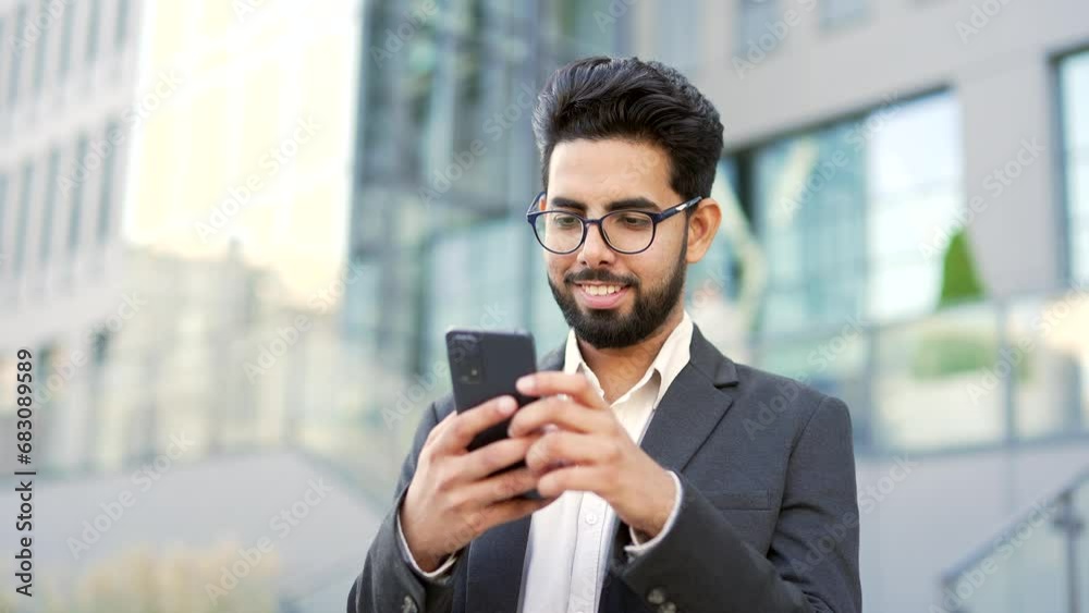 Smiling young businessman in formal suit is using a smartphone standing on street near an office building. Happy entrepreneur browsing the phone, checking email messages or chatting online. Close up Smiling young businessman in formal suit is using a smartphone standing on street near an office building. Happy entrepreneur browsing the phone, checking email messages or chatting online. Close up