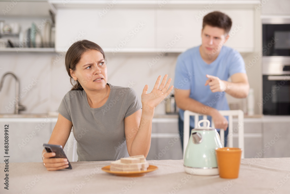 Offended upset young girl sitting at home kitchen table, brushing off discontented boyfriend standing behind and reprimanding her