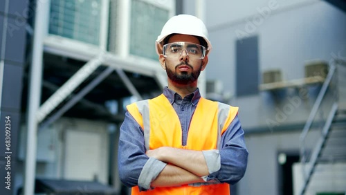 Portrait of confident professional engineer wearing safety helmet, protective glasses and vest standing at factory with crossed arms. Headshot of serious industry manager in uniform looking at camera photo
