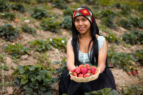 Mujer campesina bastante joven con diadema tejida sosteniendo un tazón lleno de frutas y comiendo fresas maduras en el campo después de la cosecha