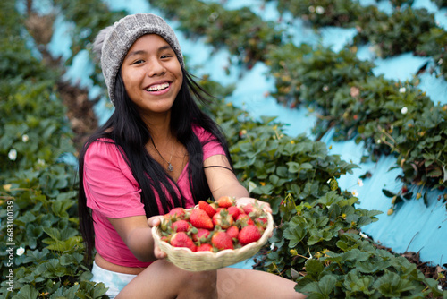 Mujer turista bastante joven con diadema tejida sosteniendo un tazón lleno de frutas y comiendo fresas maduras en el campo después de cosechar, producción de fresas. photo