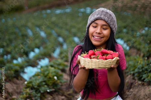 Mujer turista bastante joven con diadema tejida sosteniendo un tazón lleno de frutas y comiendo fresas maduras en el campo después de cosechar, producción de fresas.