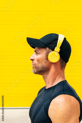 Closeup side view of serene mid adult male athlete in black cap and sportswear looking away while listening music through headphones against yellow background photo