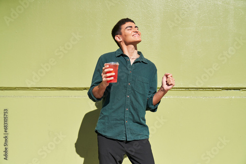 Canvas Print Young man with red iced drink outdoors by green wall