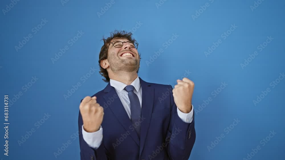 Joyful young man in business suit celebrating his win with victorious gesture, eyes filled with triumph, arms uplifted in happiness against isolated blue background. portrait of a successful winner. Joyful young man in business suit celebrating his win with victorious gesture, eyes filled with triumph, arms uplifted in happiness against isolated blue background. portrait of a successful winner.