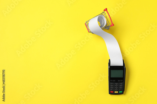 Payment terminal with thermal paper for receipt and small shopping cart on yellow background, flat lay. Space for text photo