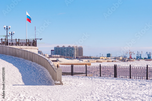 A large Russian flag on the snow-covered embankment of the Amur River on a sunny winter day. Granite coastal complex in the city of Blagoveshchensk, Russia. River port and construction cranes photo