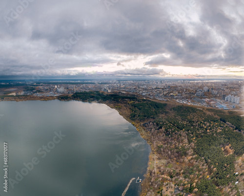 Autumn forest on lake shore at sunset and city on horizon, aerial view