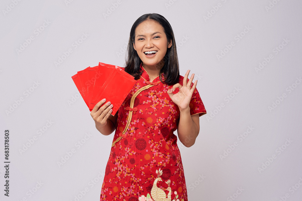 Smiling young Asian woman wearing a traditional cheongsam qipao dress holding angpao or a red packet of monetary gifts and showing okay sign isolated on white background. Happy Chinese New Year Smiling young Asian woman wearing a traditional cheongsam qipao dress holding angpao or a red packet of monetary gifts and showing okay sign isolated on white background. Happy Chinese New Year