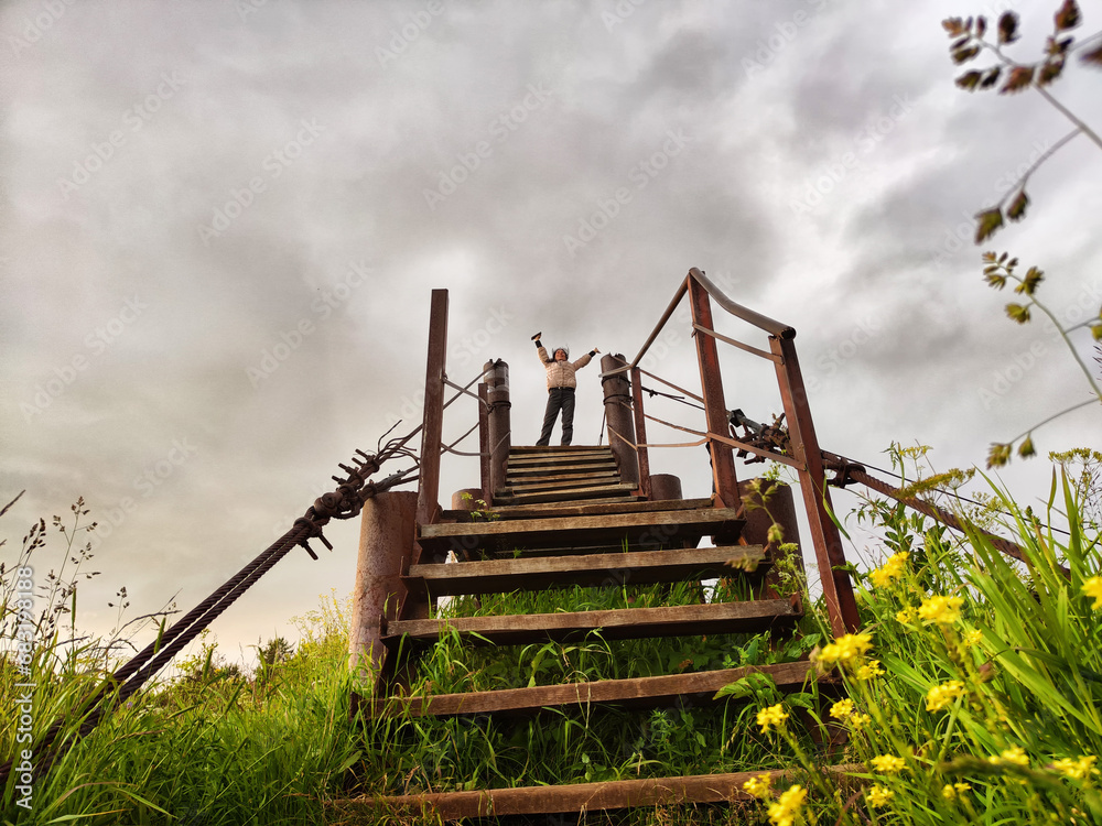 High, old, rusty iron staircase and tiny statue of girl or woman at the top against backdrop of gray, gloomy, thunderous clouds. Concept of an unusual, fantastic journey, surrealism, a strange world High, old, rusty iron staircase and tiny statue of girl or woman at the top against backdrop of gray, gloomy, thunderous clouds. Concept of an unusual, fantastic journey, surrealism, a strange world