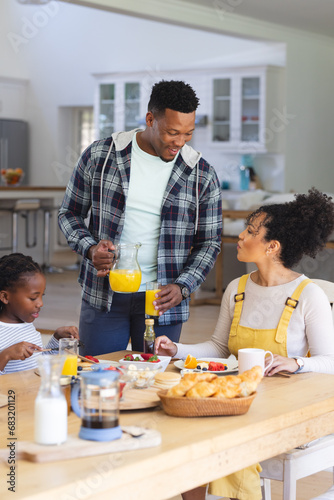 Happy african american man pouring orange juice to glass at table at home, copy space
