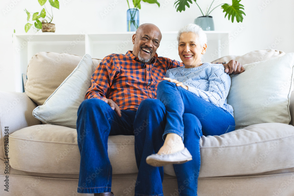 Happy diverse senior couple sitting on sofa and embracing in sunny living room
