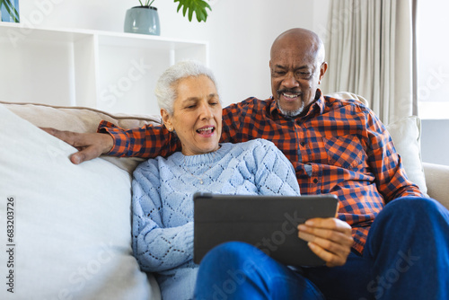 Diverse senior couple sitting on sofa, talking, embracing and using tablet in sunny living room