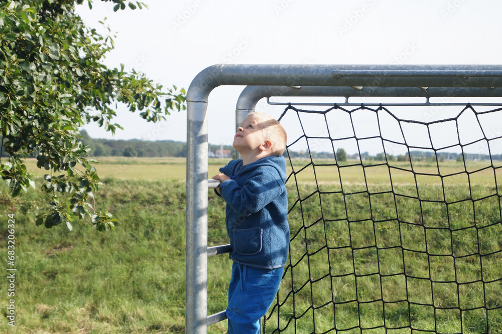 Small boy in blue jacket standing at soccer goals against the backdrop of open field, holding on to the goal frame and looking up. Children's sports games and Early skill development